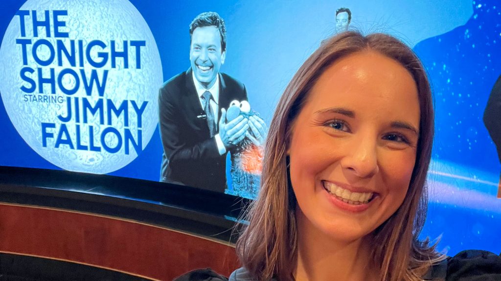 Leigh Baldwin taking a Selfie in front of a large blue banner showing Jimmy Fallon and Elmo next to the logo of "The Tonight Show Staring Jimmy Fallon"